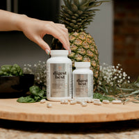 Two large bottles of Digest Best Digestive enzyme. Daily Digestive Enzyme. Decrease bloating and gas. Heartburn relief. Enhance liver health. NUsisters. 60 Capsules. 120 capsules. Capsules on the counter. Pineapple, spinach, and flowers in the background. A woman is placing her hand on one of the bottles.