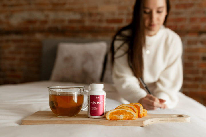 A large bottle of elever woman's multivitamin. Balance. Boost. Digestion. Energy. Renew. Refresh. 60 capsules. With a tea and oranges and a women writing in a book in the background.