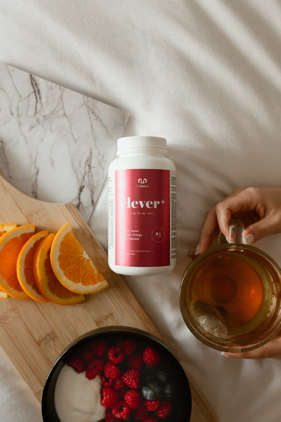 A large bottle of elever woman's multivitamin. Balance. Boost. Digestion. Energy. Renew. Refresh. 120 capsules. A woman is holding a tea. There are also oranges and a bowl of yogurt and berries.