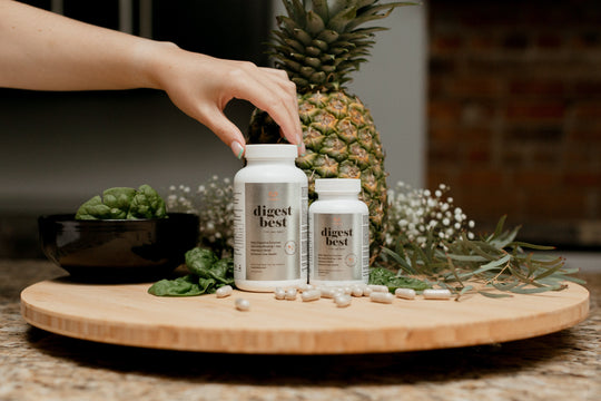 Two large bottles of Digest Best Digestive enzyme. Daily Digestive Enzyme. Decrease bloating and gas. Heartburn relief. Enhance liver health. NUsisters. 60 Capsules. 120 capsules. Capsules on the counter. Pineapple, spinach, and flowers in the background. A woman is placing her hand on one of the bottles.